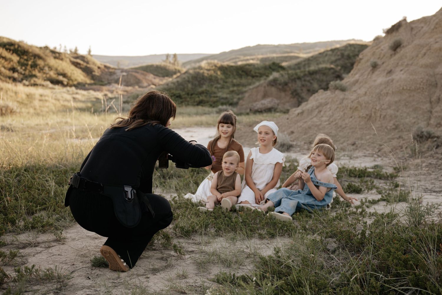 Woman crouching to photograph group of children sitting outdoors in sandy landscape at dusk.