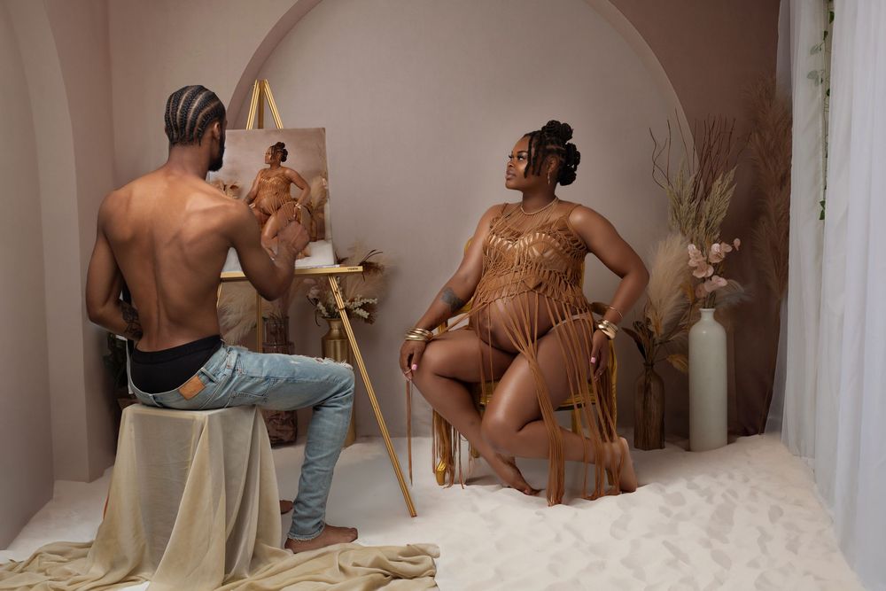 A shirtless man paints a portrait of a pregnant woman in a gold bodysuit seated on a chair in a studio.