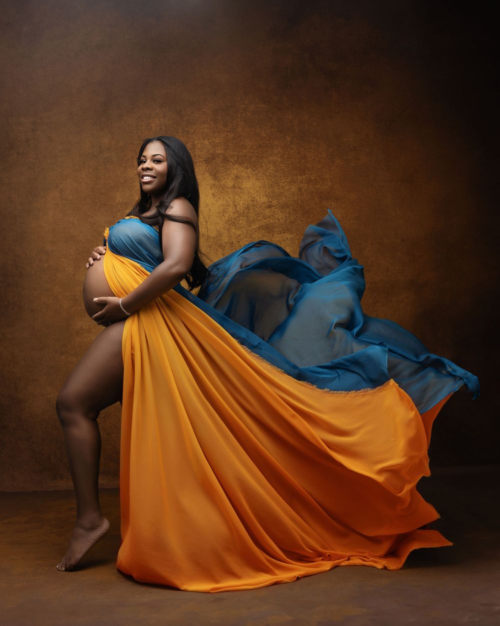 Pregnant woman in flowing teal and orange gown cradling baby bump in a stunning studio maternity portrait.