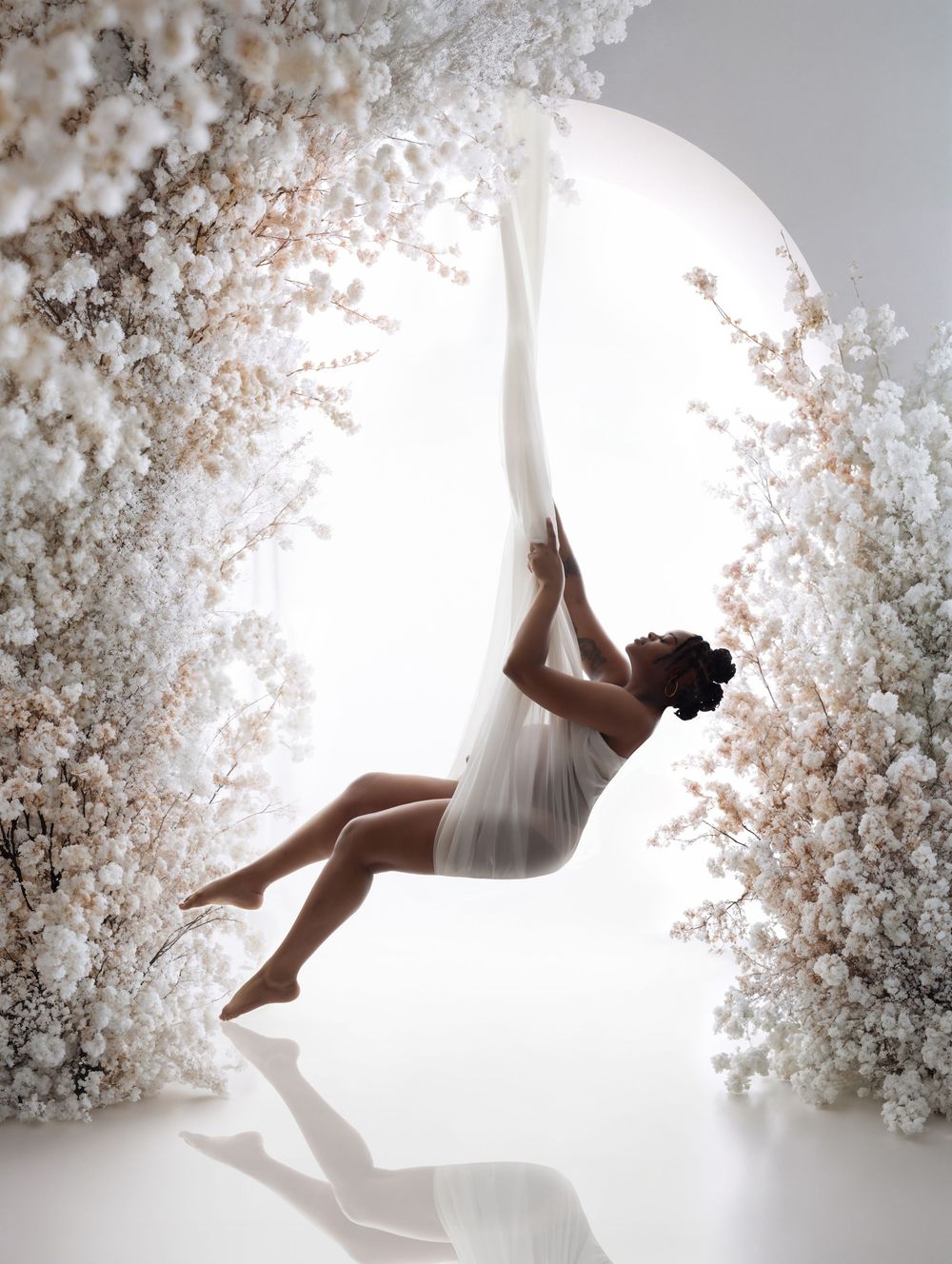 A dancer in white draping fabric swings gracefully amid lush white floral arches in a dreamy, ethereal studio setting.