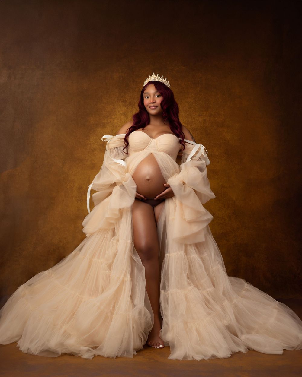 Pregnant woman in elegant champagne tulle gown and tiara poses gracefully in a studio maternity portrait.