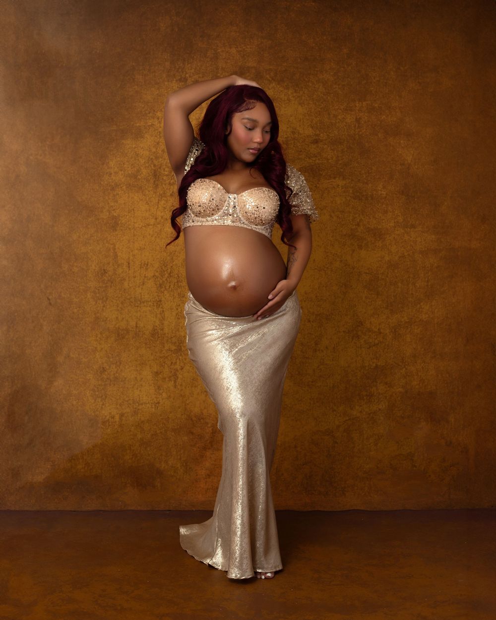 Pregnant woman in gold lace bralette and metallic skirt poses for maternity photo against golden backdrop.