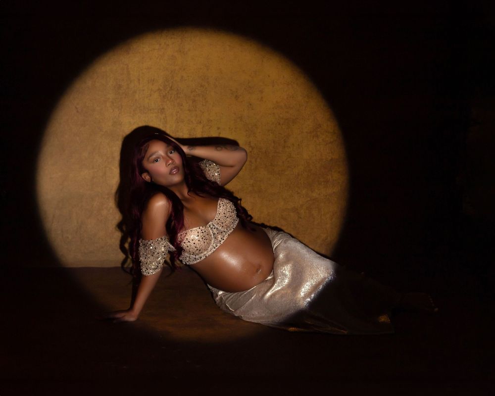 Pregnant woman in gold sequin top poses gracefully under spotlight against dark background.