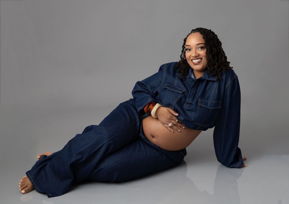 Pregnant woman in denim outfit posing on studio floor, showcasing baby bump in stylish maternity photoshoot.