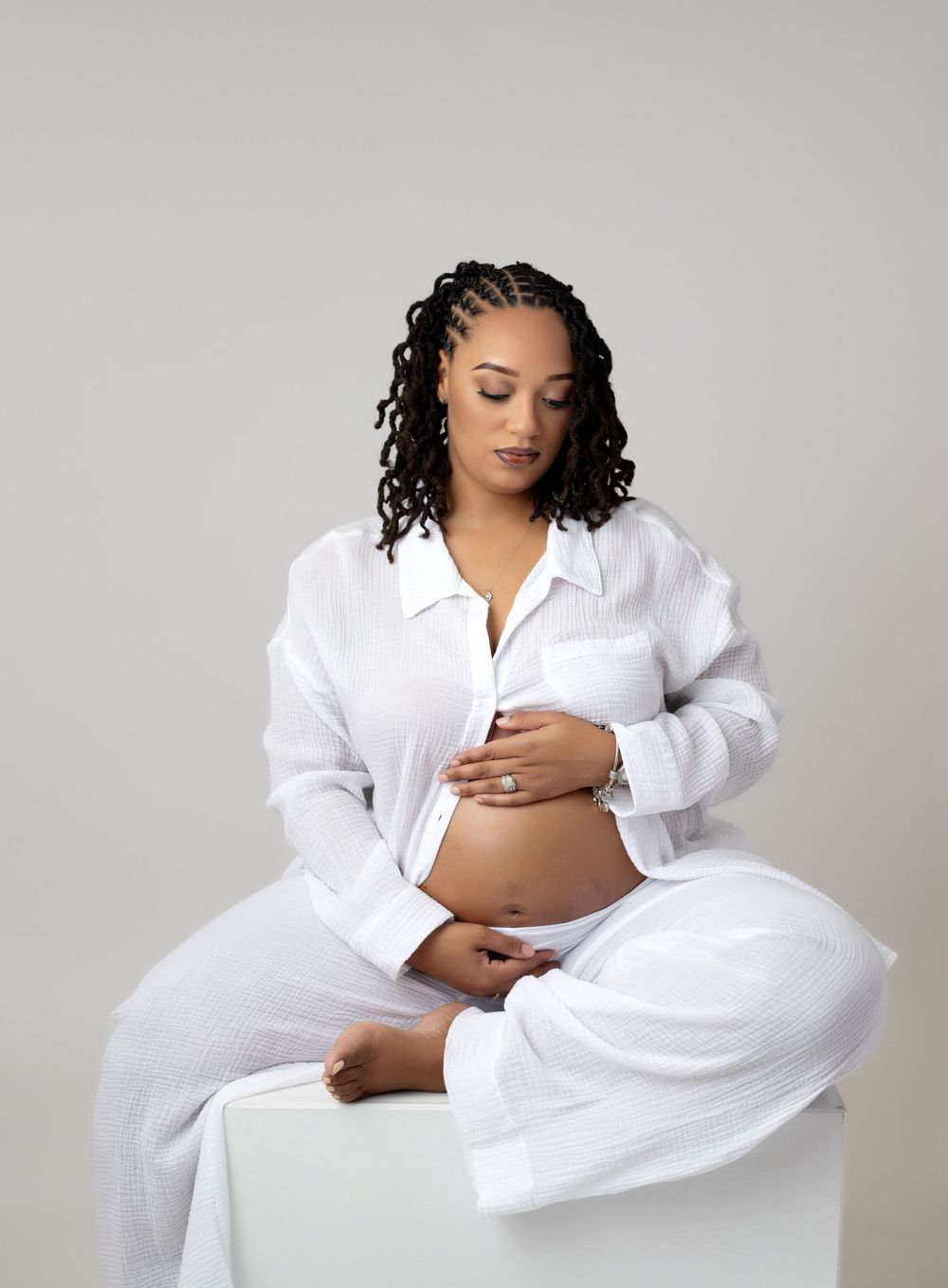 Pregnant woman in white linen outfit sits gracefully on white cube, cradling her baby bump in a serene studio portrait.