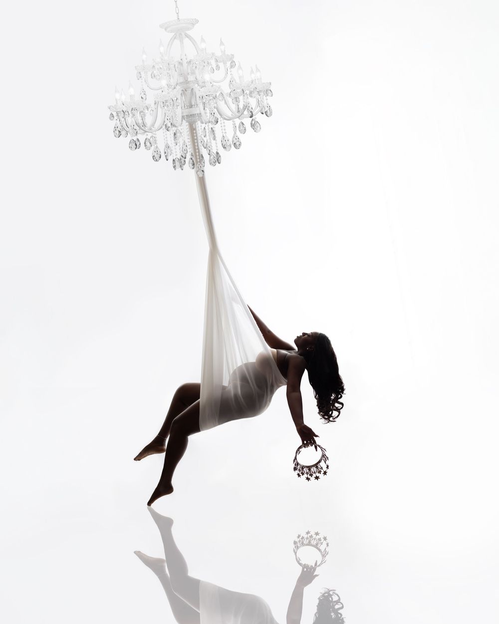 Silhouette of a woman suspended from a white chandelier using aerial silk, holding a jeweled hoop, reflected on glossy floor.