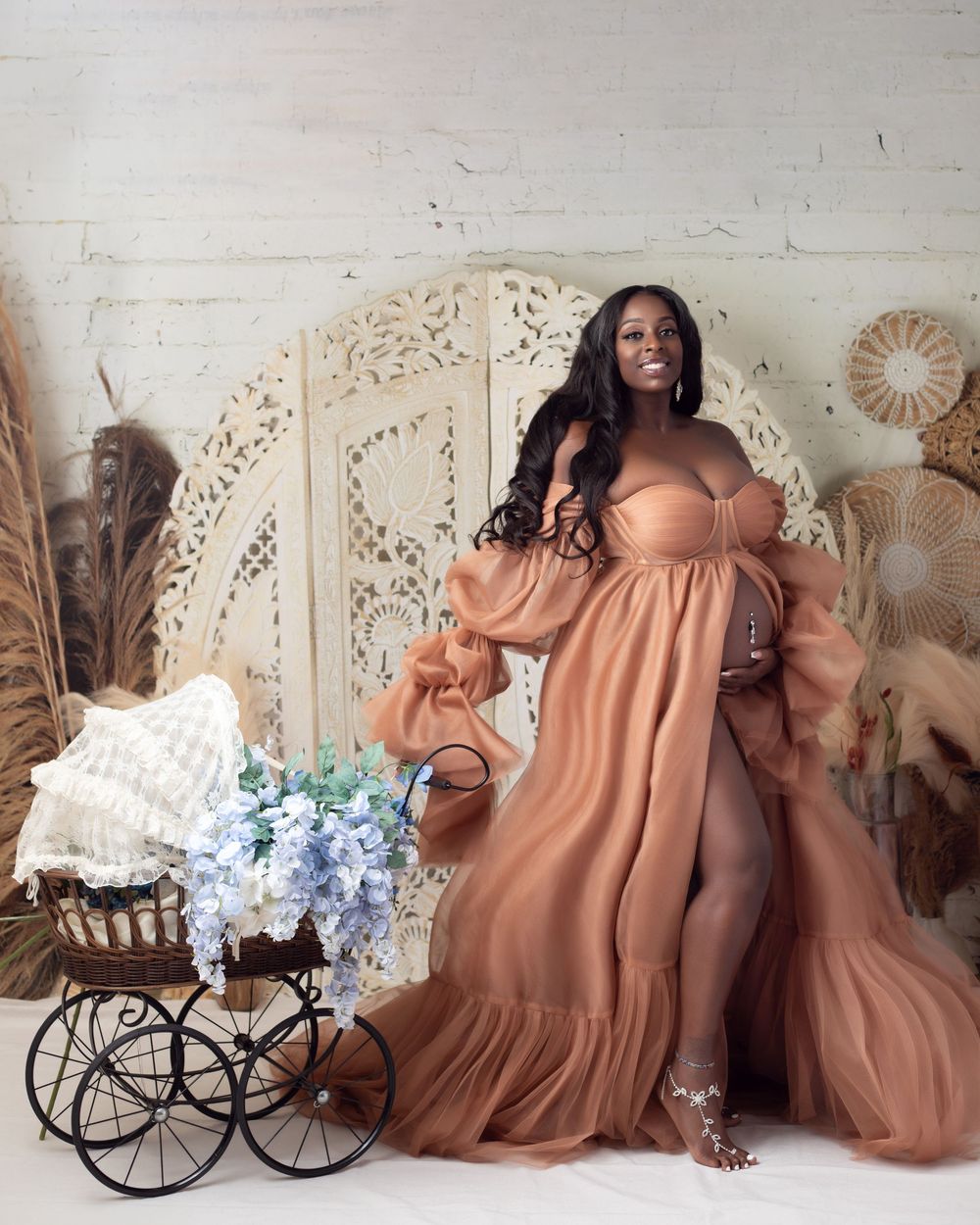 Pregnant woman in elegant rust tulle maternity gown poses beside vintage baby carriage with blue flowers in boho studio.