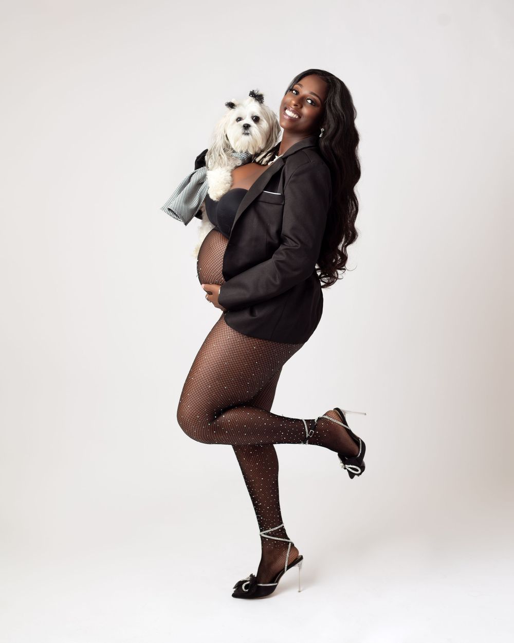 Pregnant woman in black blazer and sparkly tights holds white Maltese dog during elegant maternity photo shoot.