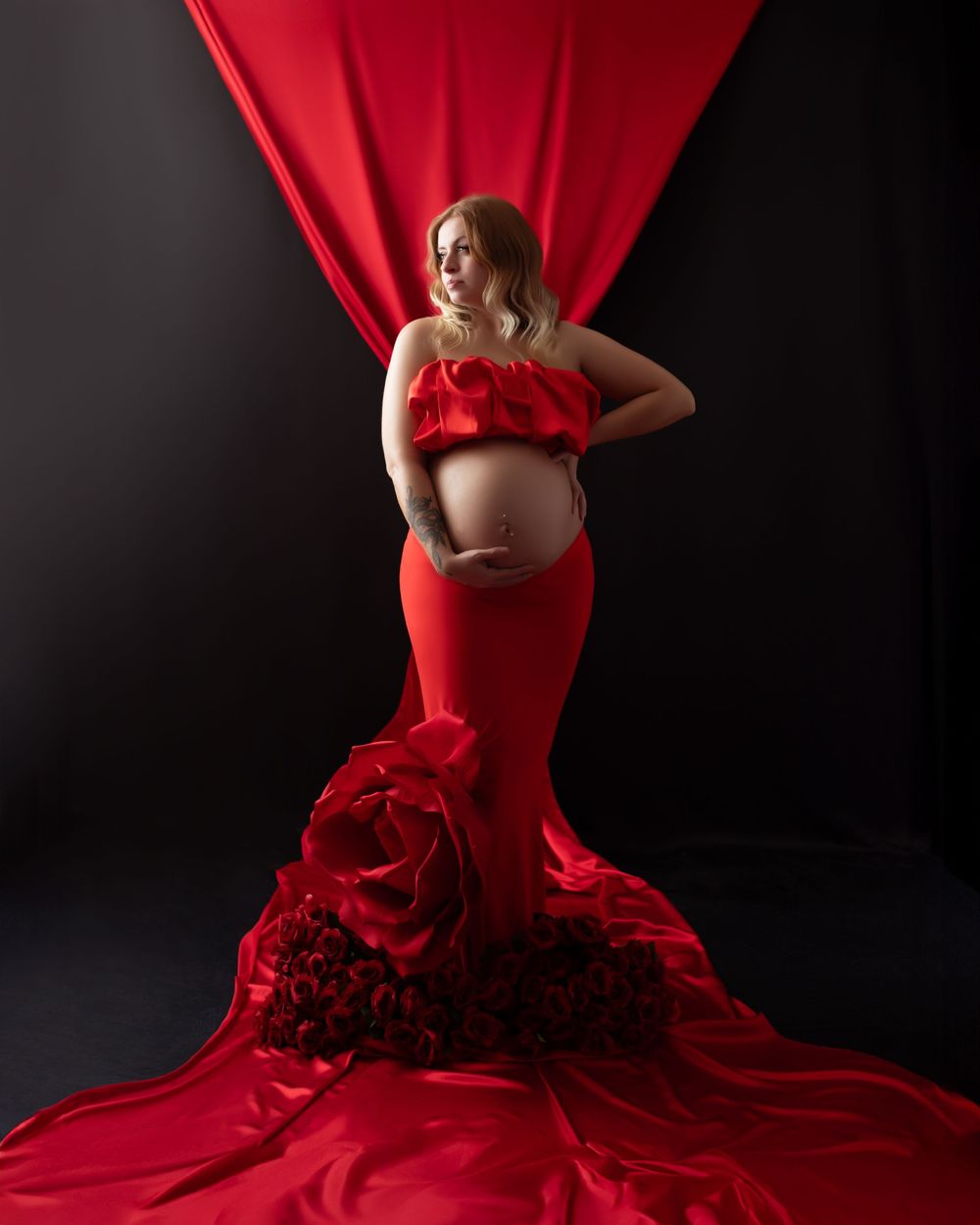 Pregnant woman in red ruffled maternity gown with floral train poses against dramatic black backdrop with red fabric drape.