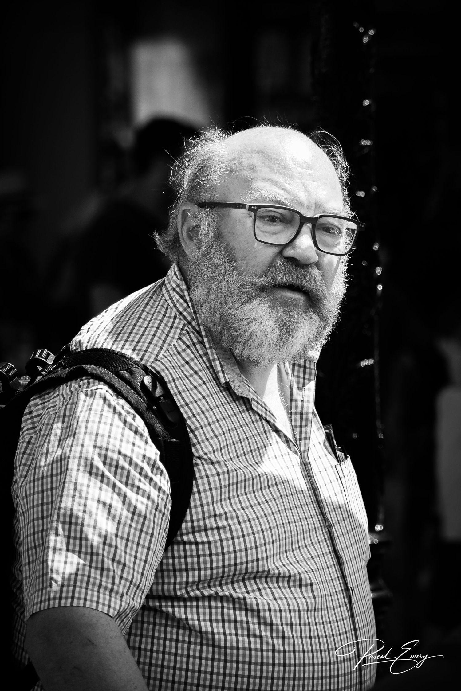 Black and white portrait of an elderly bearded man wearing glasses and a plaid shirt with a backpack.