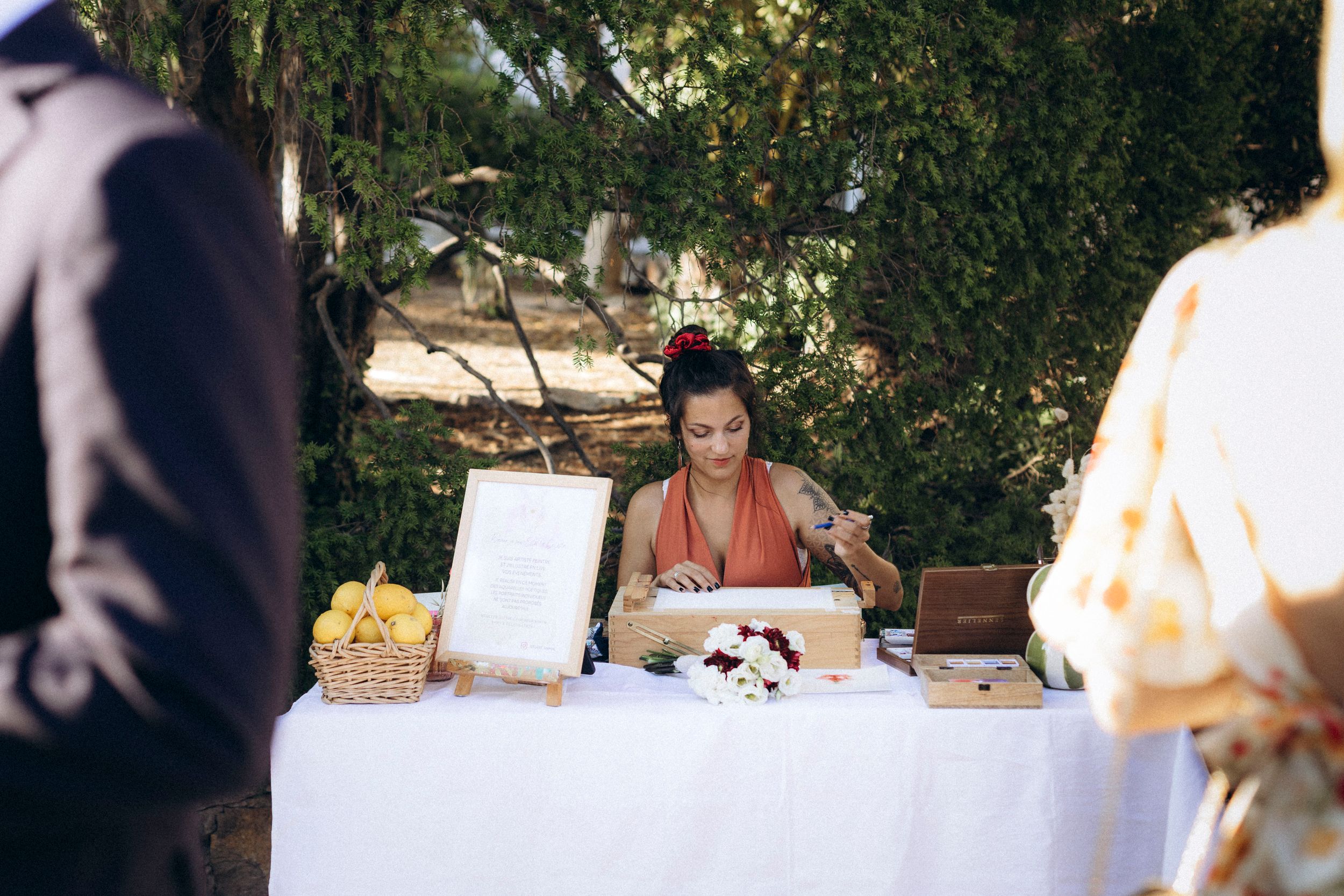 Woman in red dress writing at an outdoor wedding table with flowers, lemons, and decorative items under green trees.