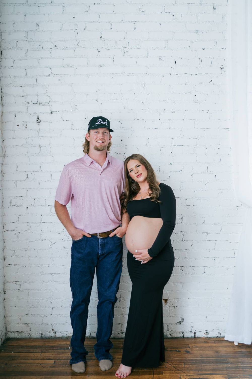 Smiling pregnant couple standing against white brick wall, man wearing Dad cap in pink polo in North Carolina
