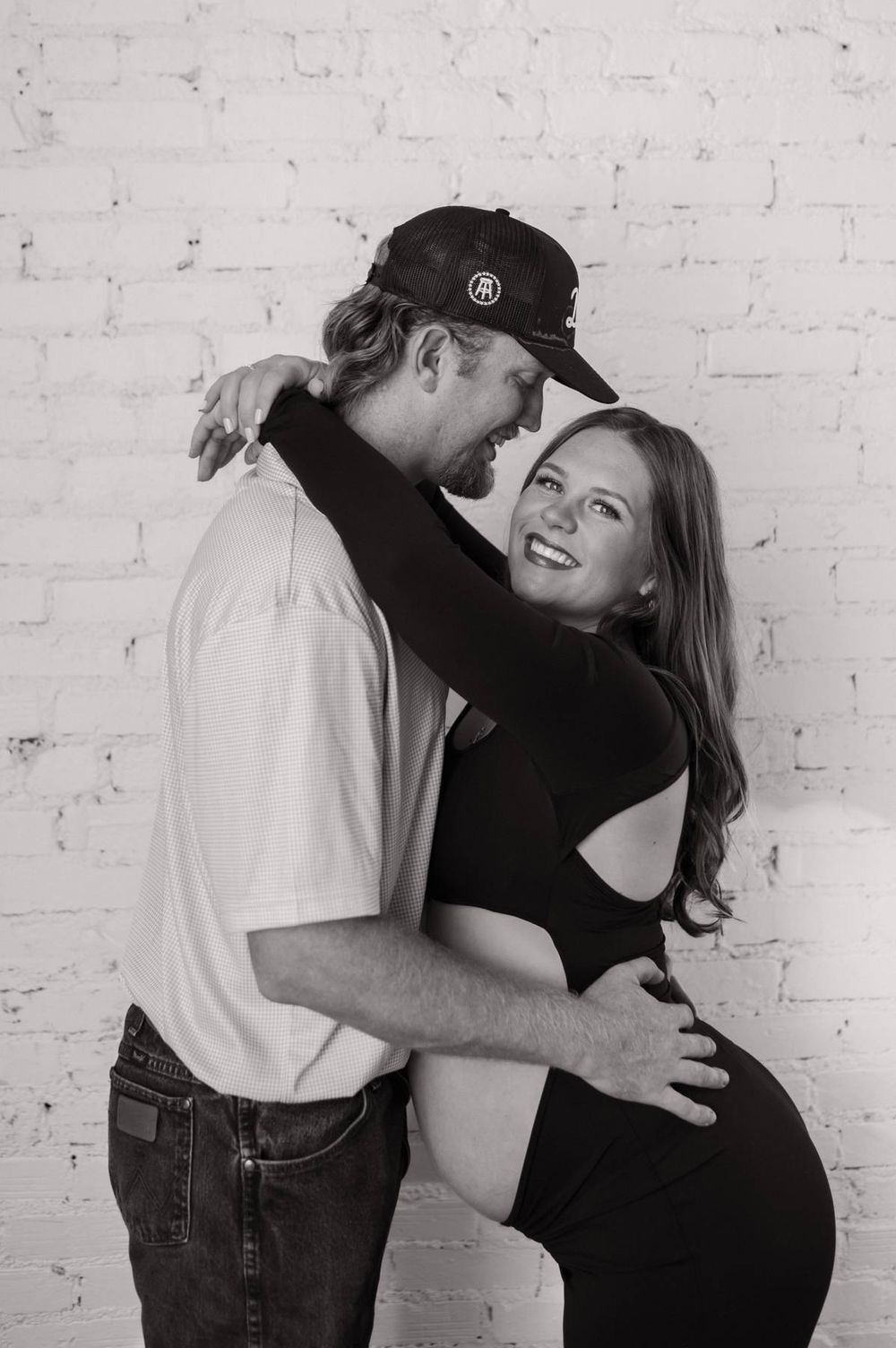 Black and white maternity photo of laughing couple embracing against white brick wall Lexington NC