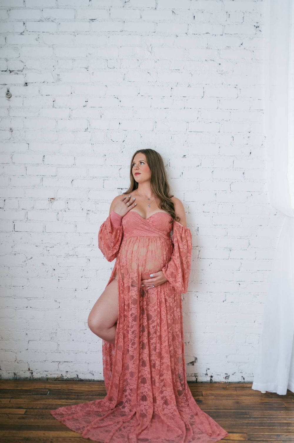 Pregnant woman in elegant pink lace maternity gown posing against white brick wall High Point NC
