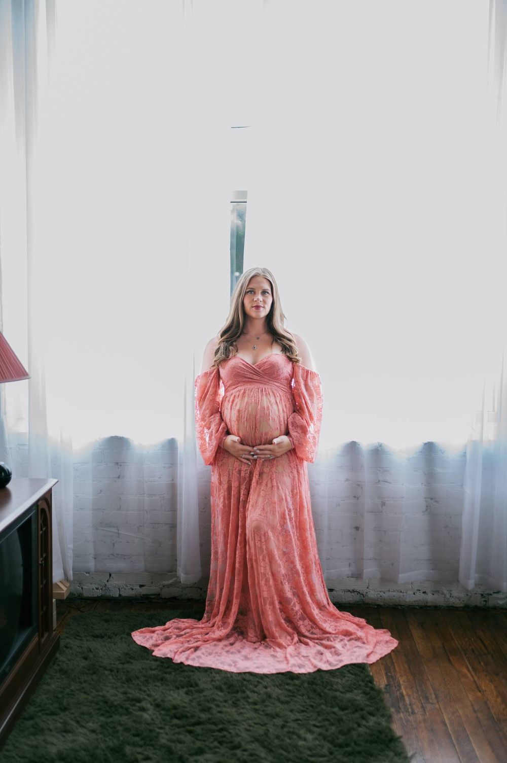 Pregnant woman in flowing pink lace gown standing before bright window in maternity portrait Greensboro NC