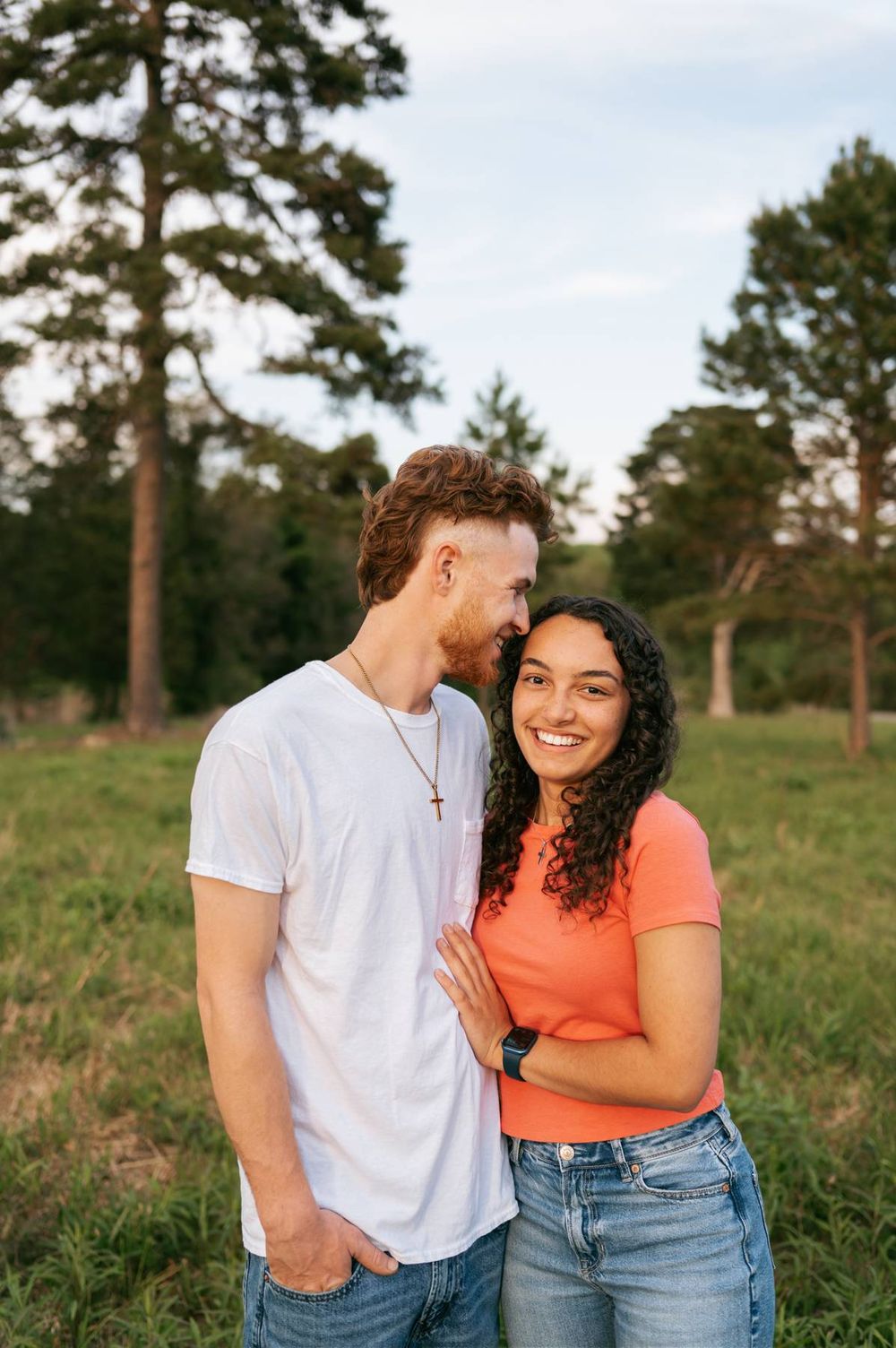 Smiling couple posing in a green meadow surrounded by tall pine trees during golden hour, couples photographer in North Carolina