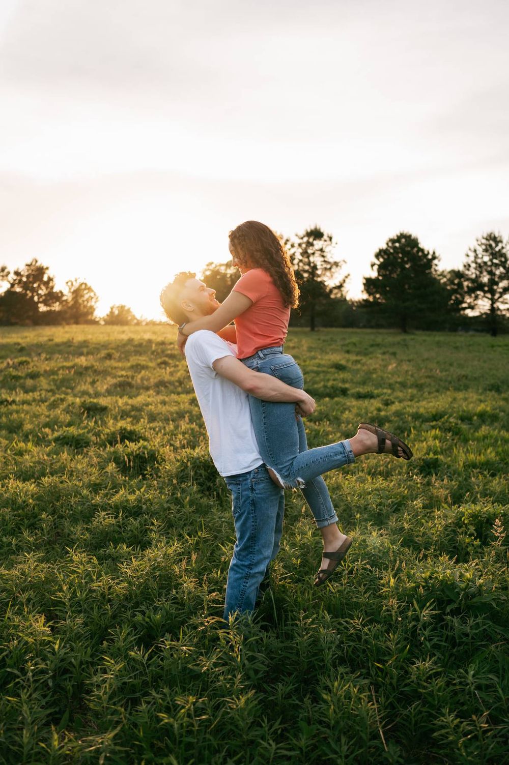 Man lifting woman in a romantic embrace in a lush green field during a glowing sunset, documentary photographer