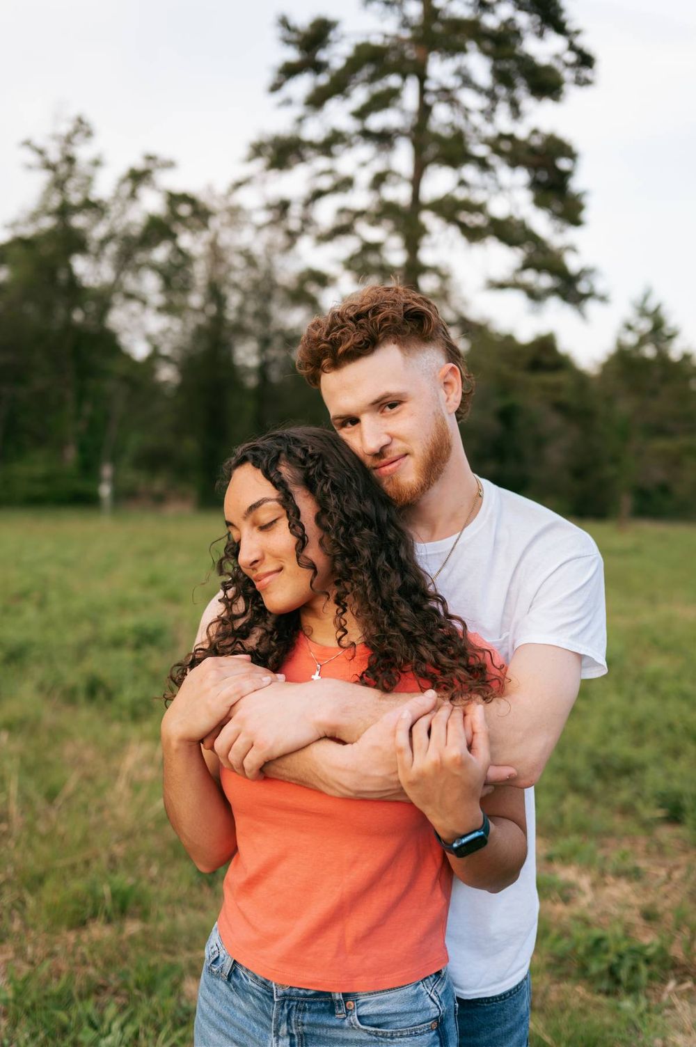 Man embracing woman from behind in a green meadow, both smiling softly in warm evening light in North Carolina