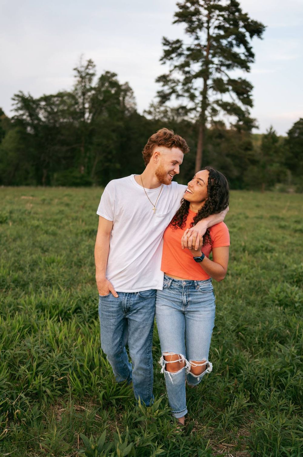Laughing couple walking together through a green field with tall pine trees in the background in North Carolina