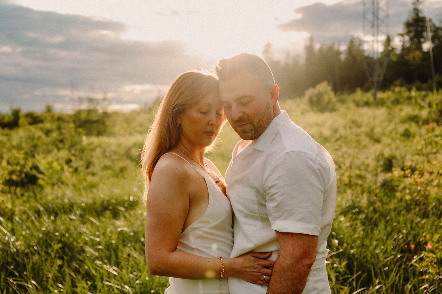Couple embracing in a sunlit meadow at golden hour, surrounded by lush green grass and trees.