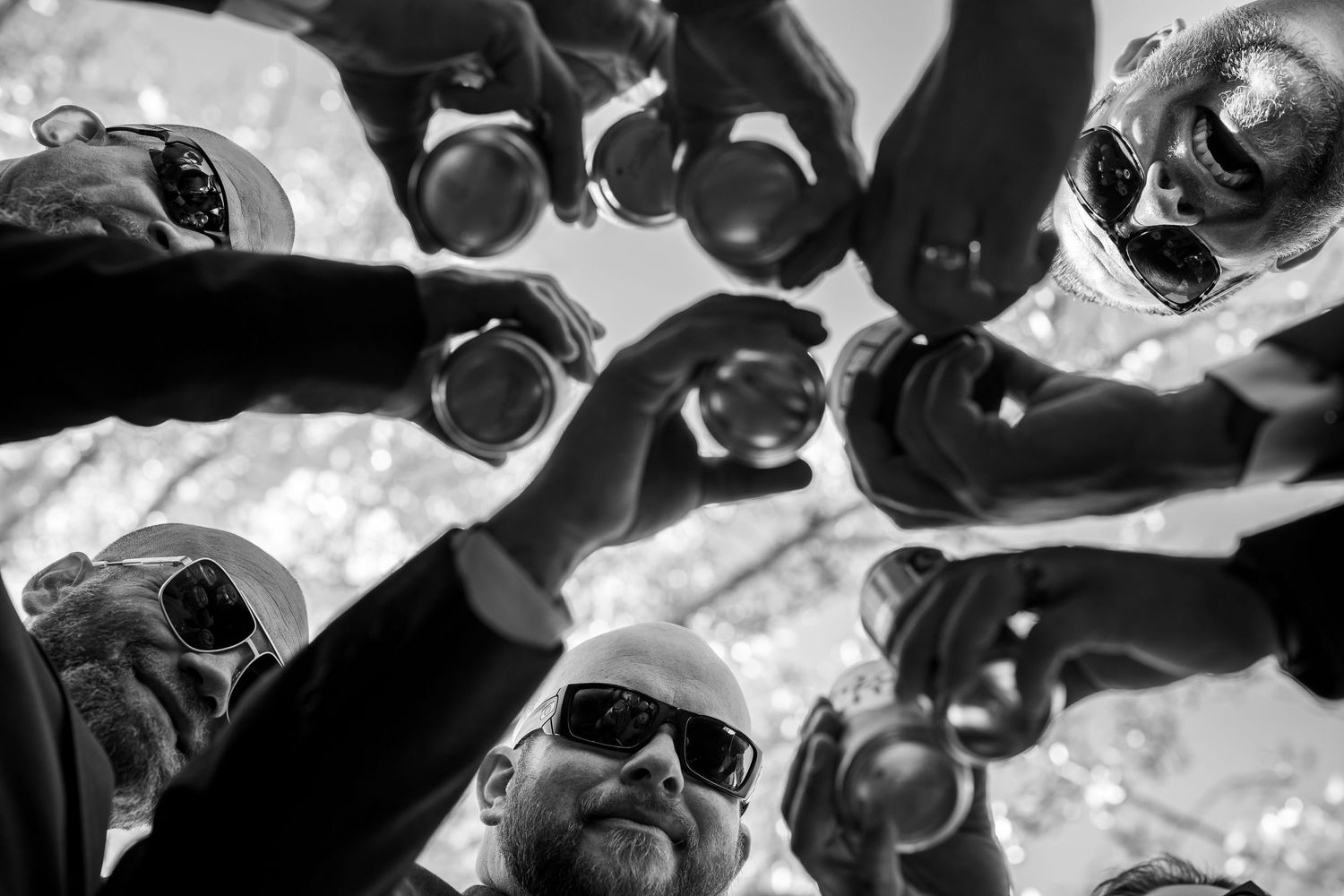 Black and white photo of friends toasting drinks in a circle, viewed from below, wearing sunglasses outdoors.