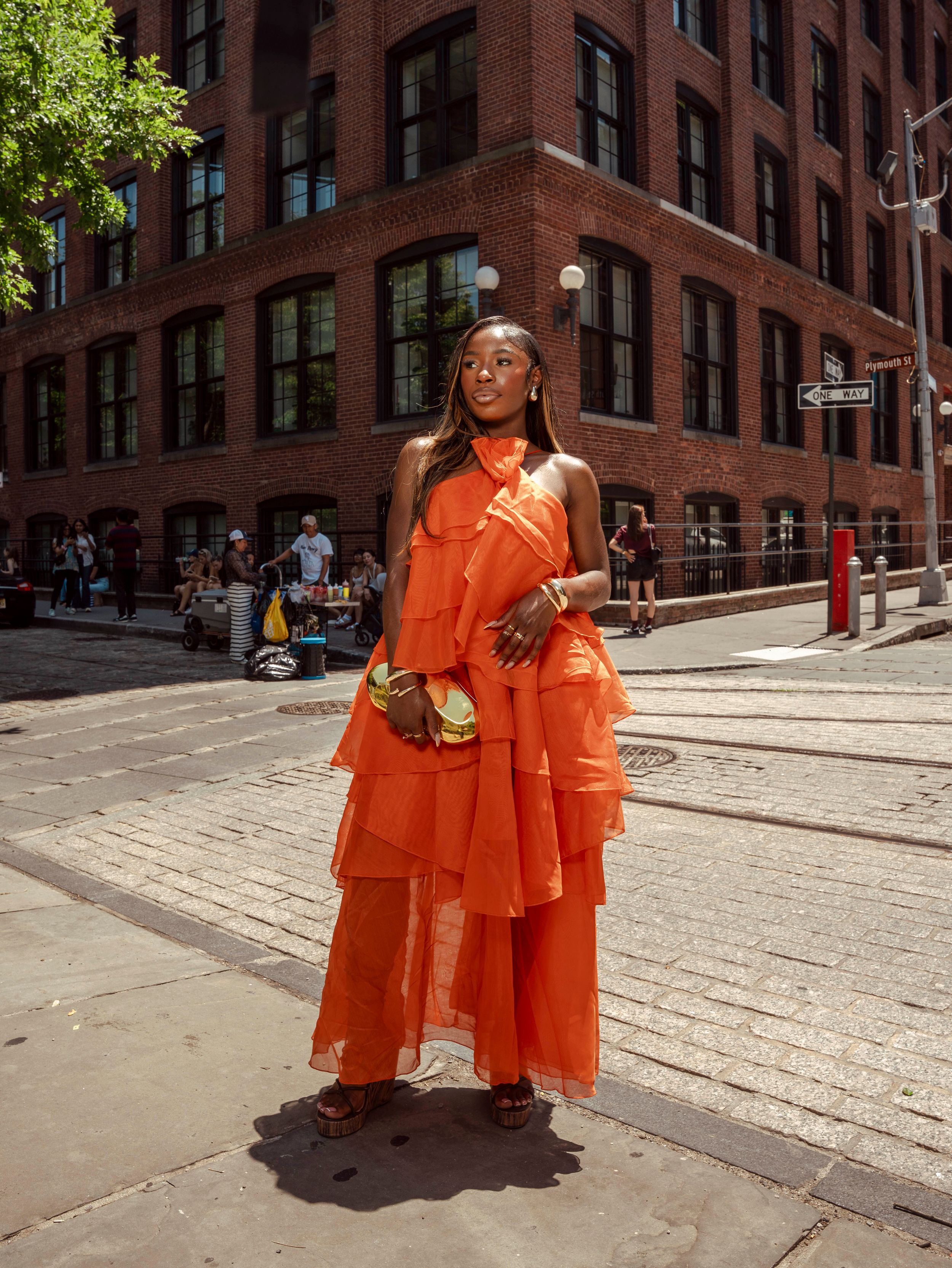 Woman in orange layered ruffle maxi dress holding clutch on cobblestone street corner NYC.