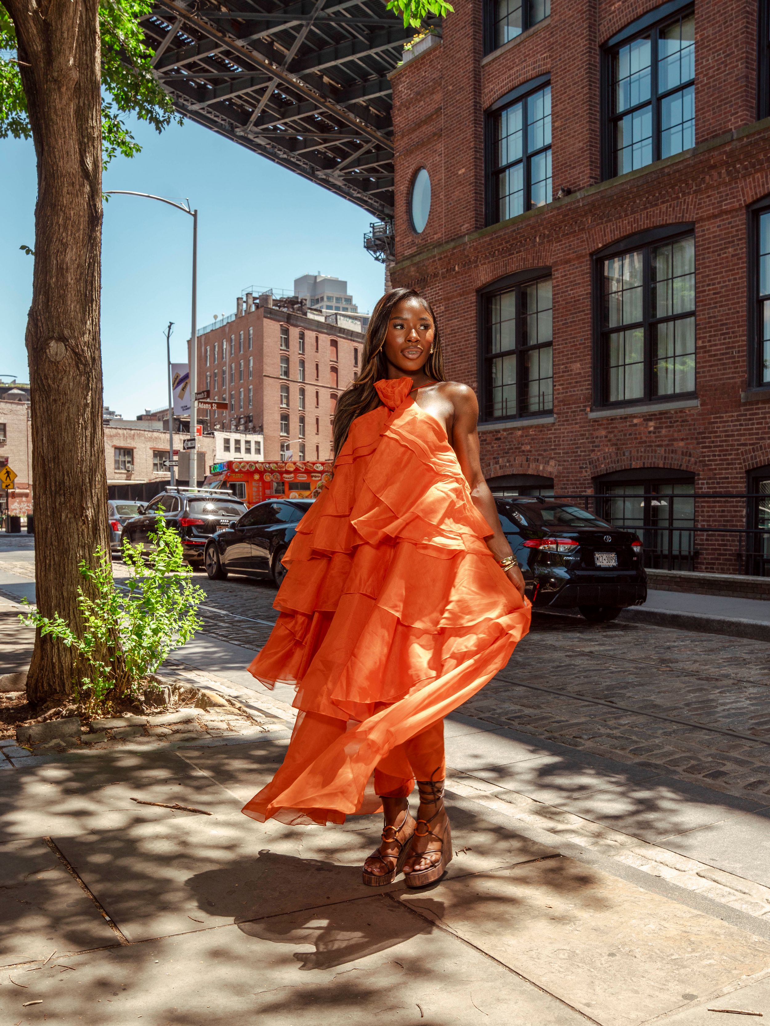 Woman twirling in vibrant orange tiered ruffle halter dress on sunny NYC sidewalk.