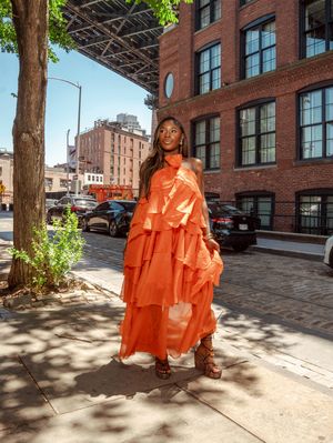Full length view of woman in flowing orange ruffled maxi dress near brick building in NYC.