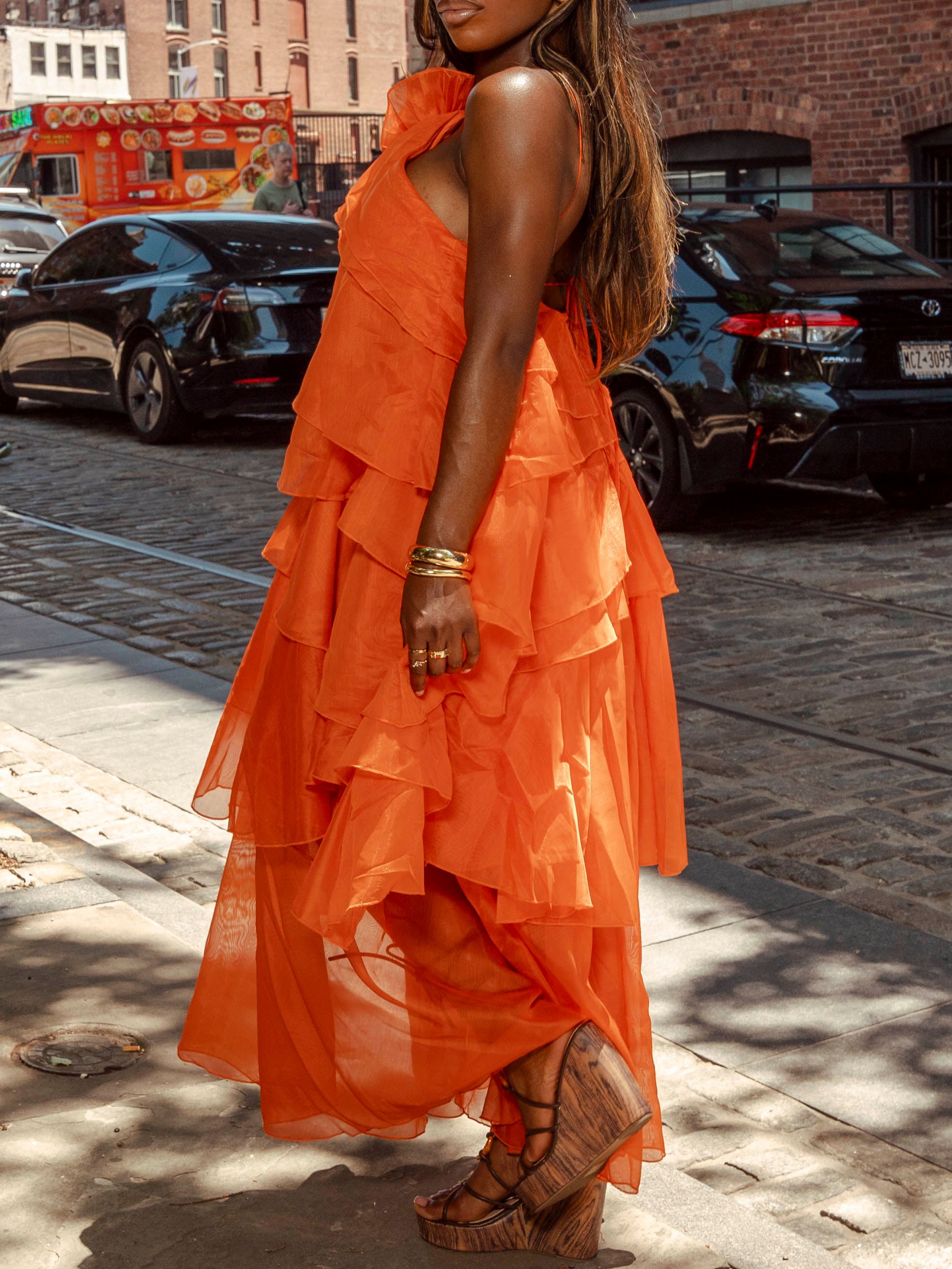 Side view of woman in bold orange tiered ruffle maxi dress on cobblestone street.