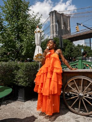 Woman in orange ruffled maxi dress near Brooklyn Bridge enjoying outdoor summer day.
