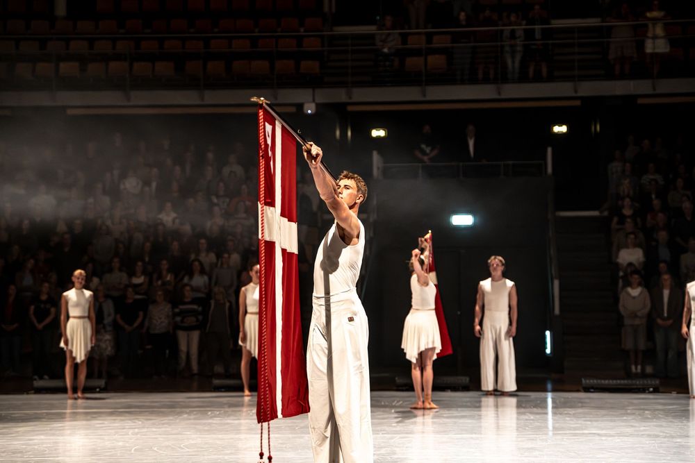 Performer in white outfit holds a Danish flag on stage with dancers in background during a theatrical performance.