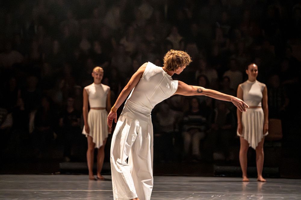 Three dancers in white costumes perform on a dimly lit stage before an audience.