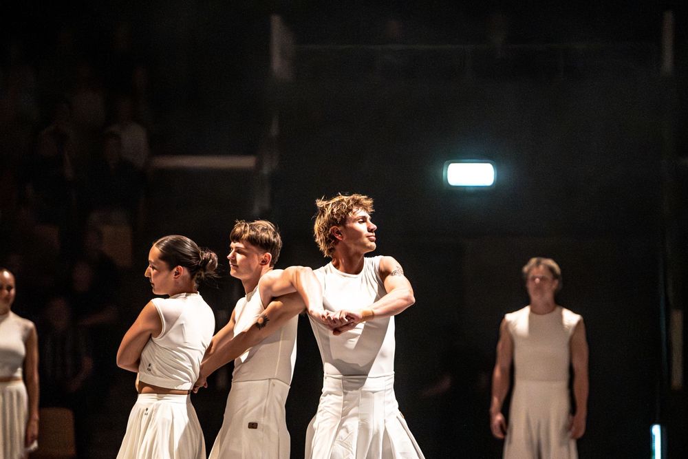 Dancers in white costumes perform a contemporary dance piece on a dark stage under bright lighting.