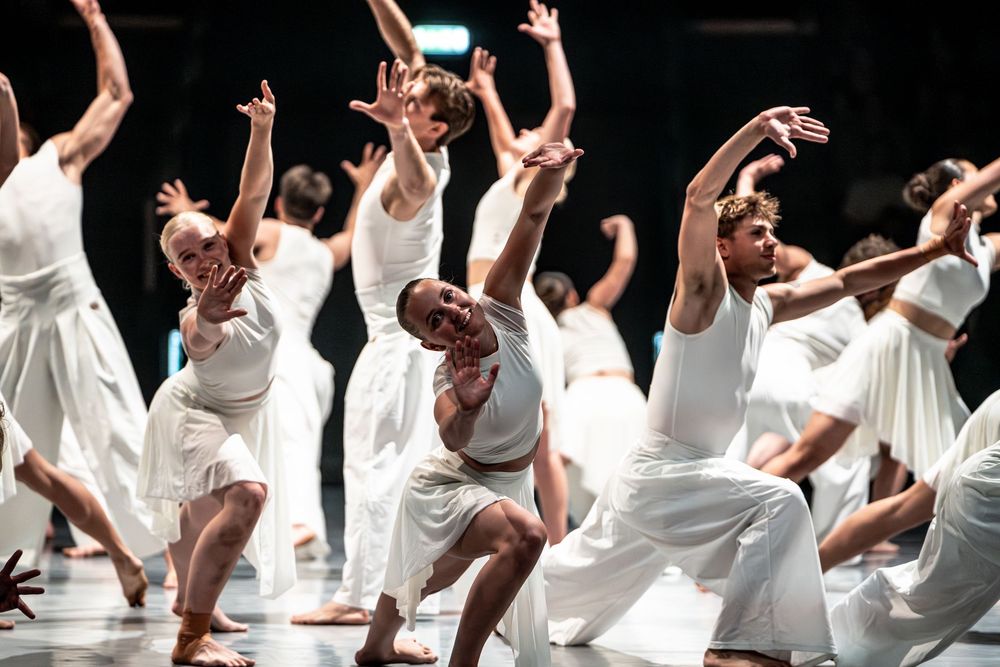Large group of contemporary dancers in white costumes performing dynamic choreography on stage.
