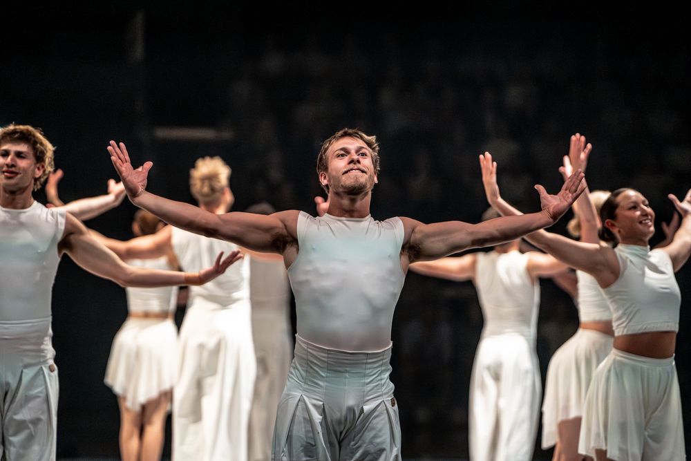Dancers in white costumes perform on stage, with a male dancer center front arms outstretched in a dramatic pose.
