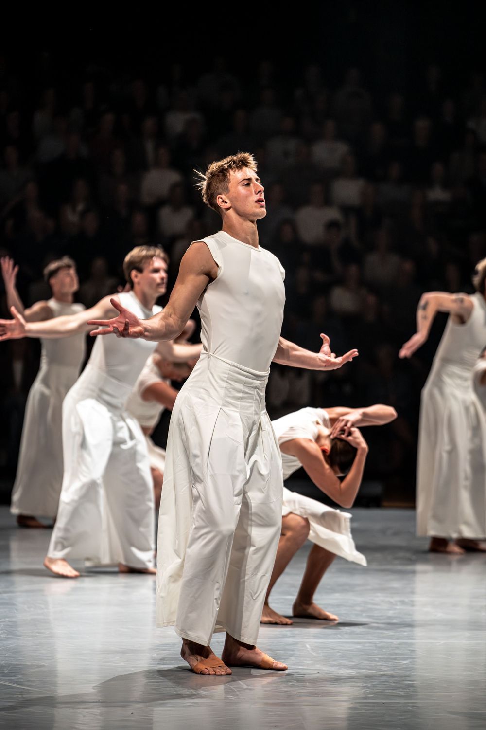 Male contemporary dancer in white costume performs expressive modern dance on stage with ensemble in background.