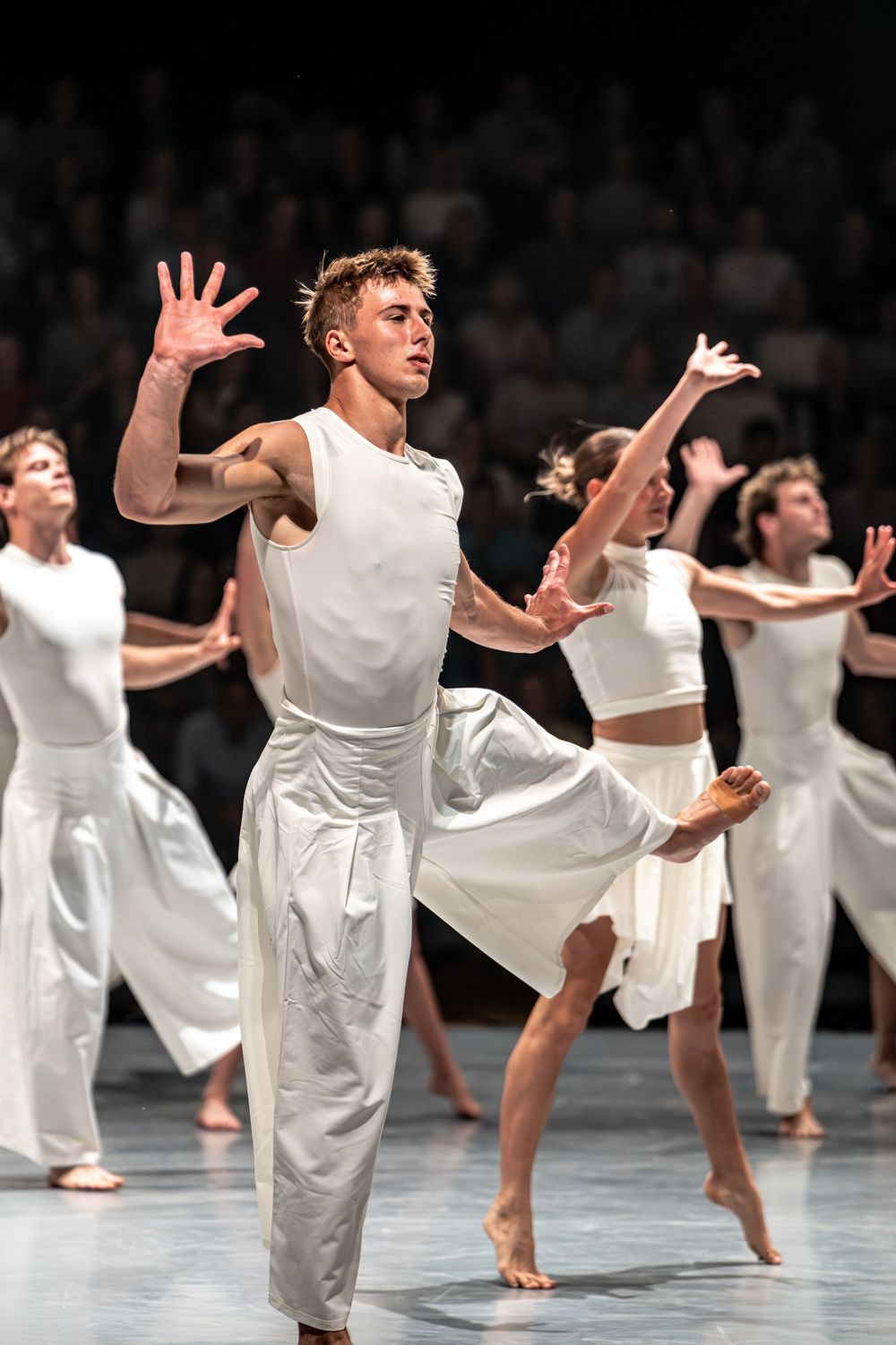 Male contemporary dancer in white leads ensemble performing expressive modern dance on stage.