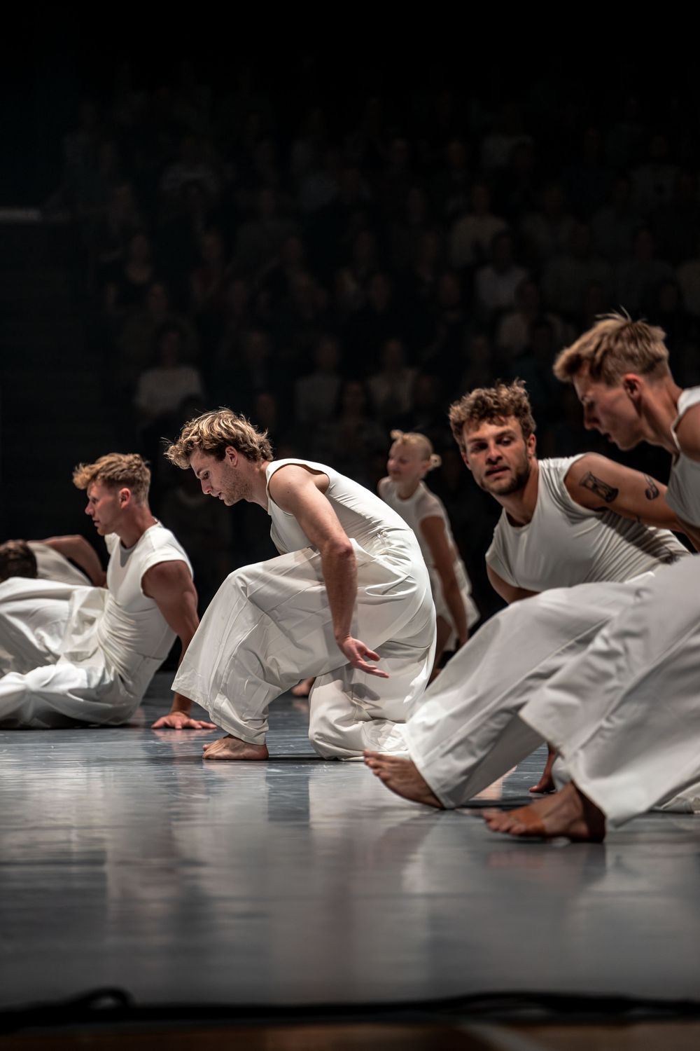 Male dancers in white costumes perform a contemporary dance routine on stage before a live audience.