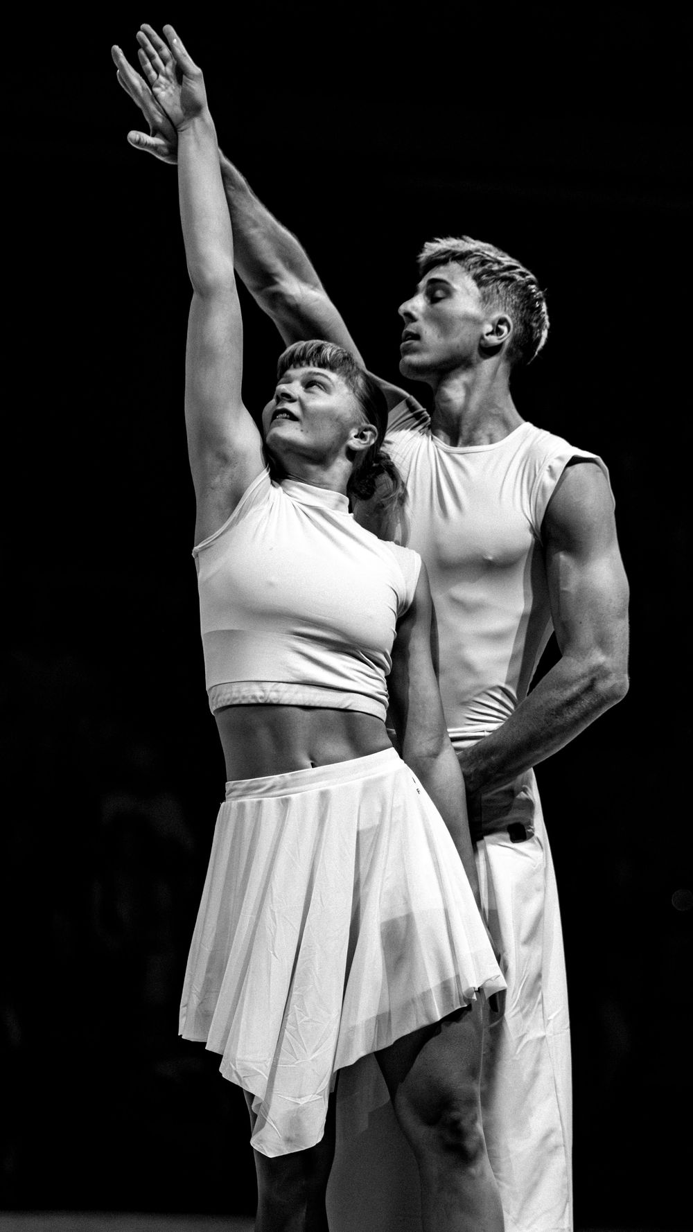 Two ballet dancers in white perform an elegant pas de deux, woman reaching upward, black background.