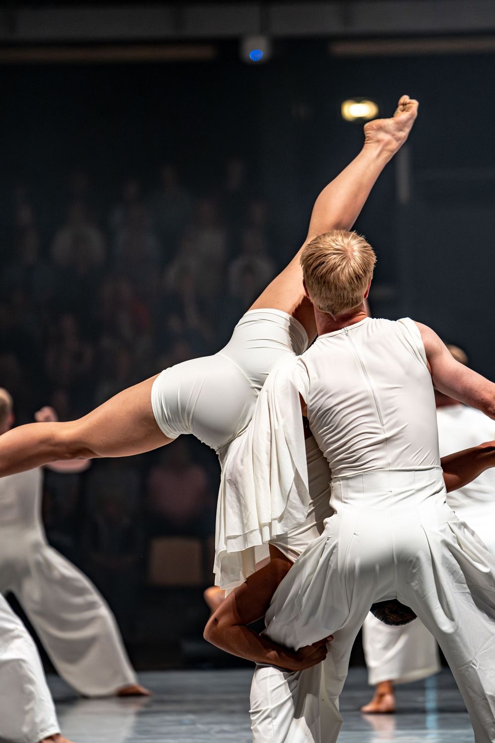 Two dancers in white costumes perform a dramatic lift on stage before an audience.