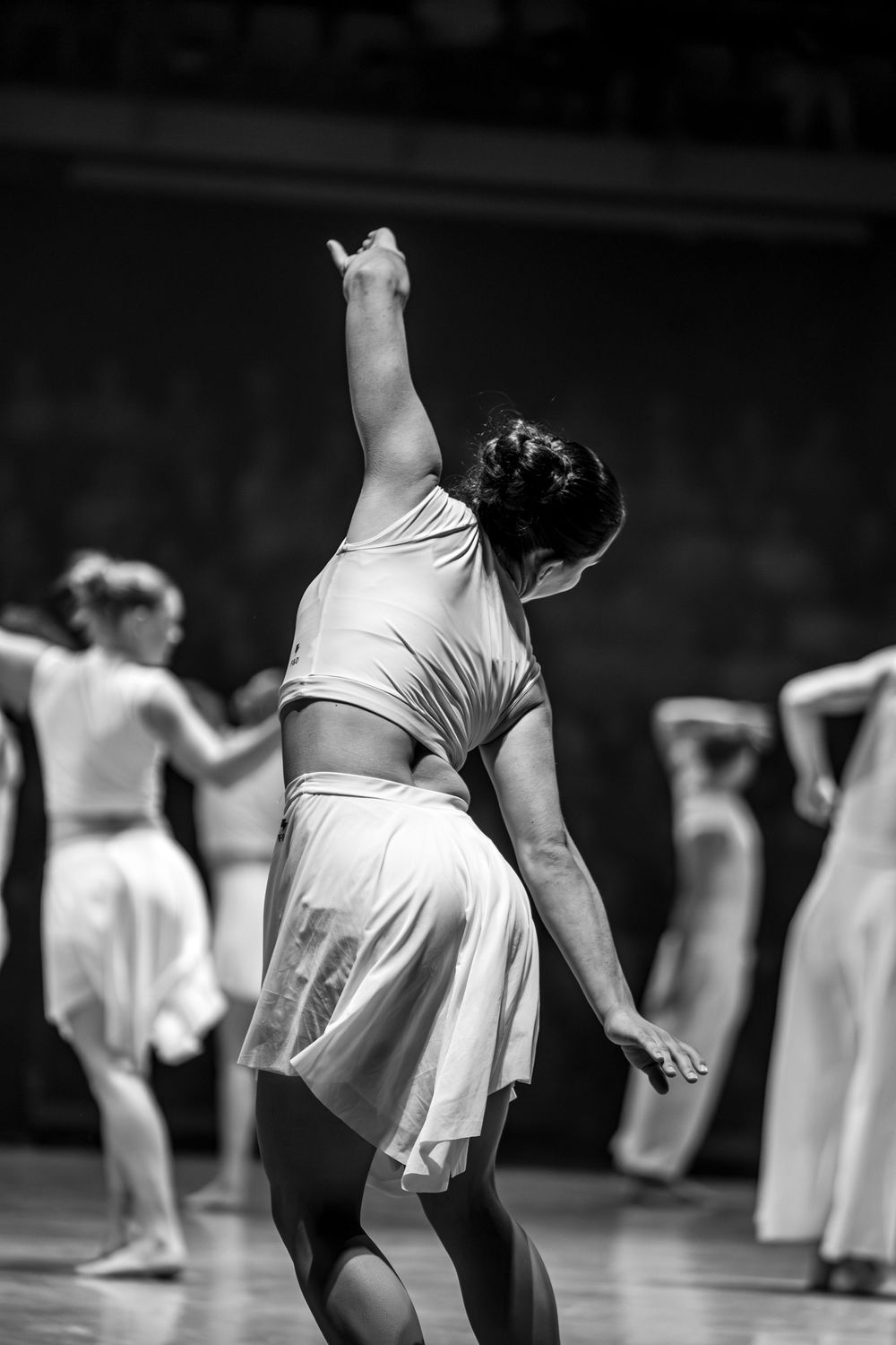 Black and white photo of a contemporary dancer in white reaching upward during a stage performance.