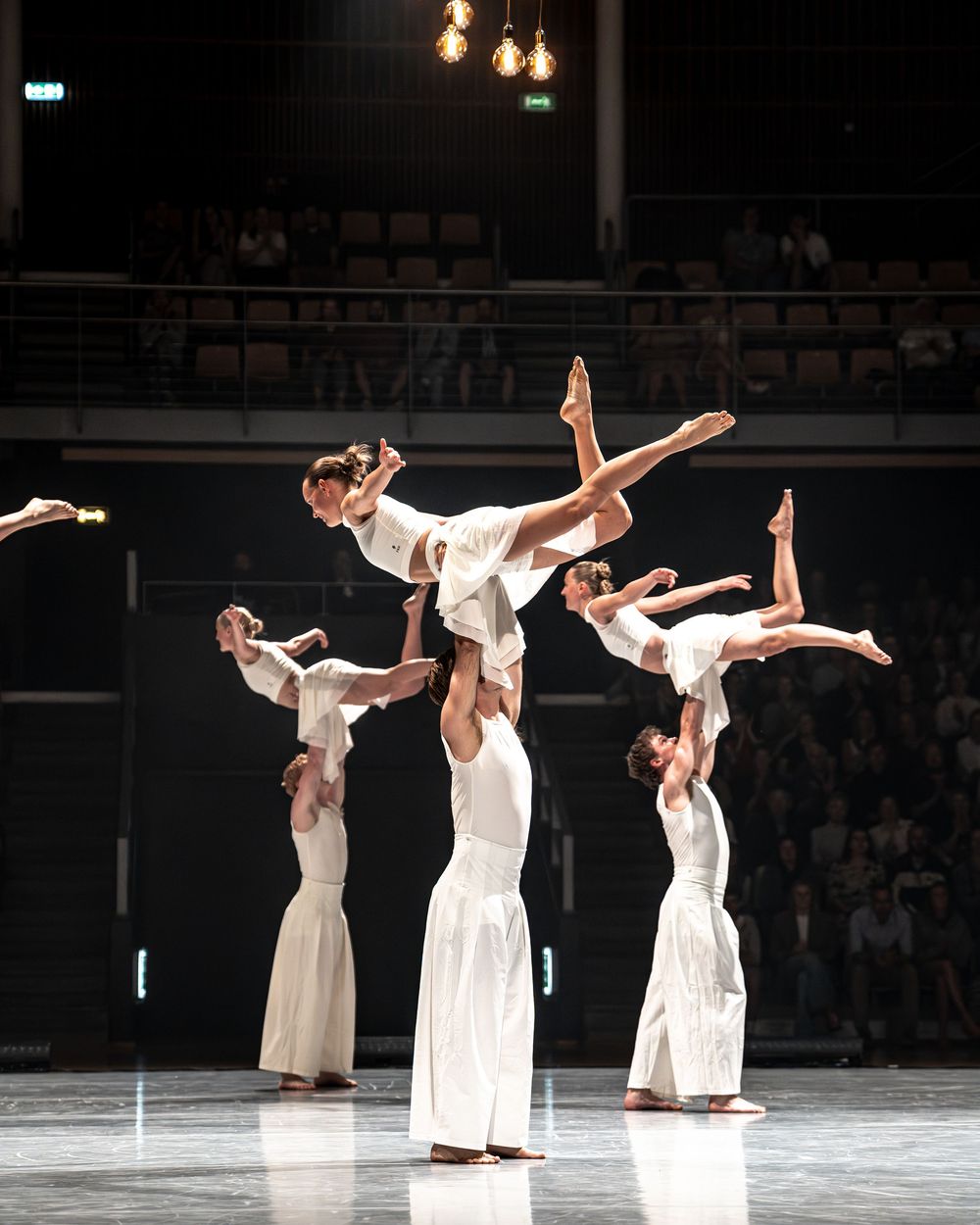 Contemporary dancers in white costumes perform acrobatic lifts on a theater stage during a modern dance performance.
