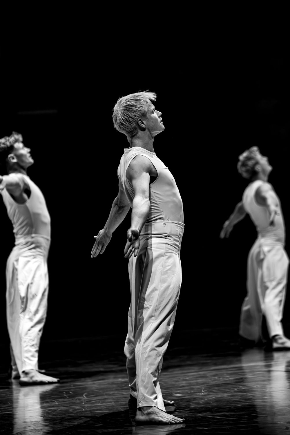 Male dancers in white costumes perform on a dark stage in black and white photo.