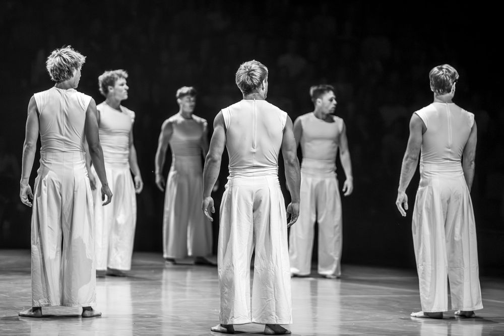 Black and white photo of male dancers in white costumes performing on stage in a modern dance production.