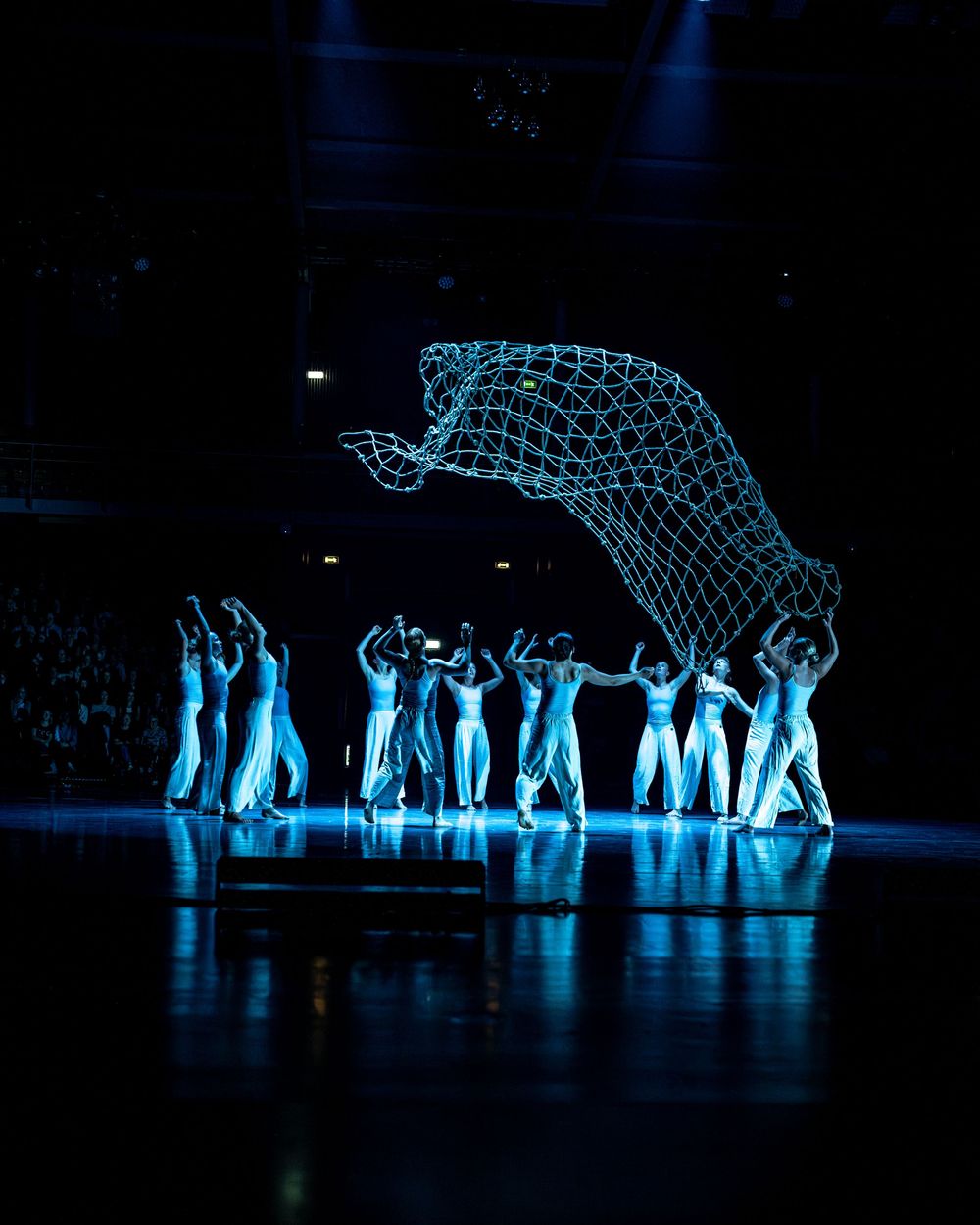 Dance ensemble in white costumes supports glowing wireframe elephant sculpture in dramatic dark stage performance.