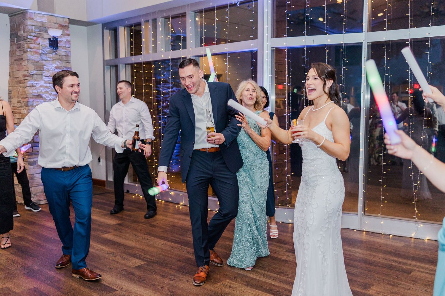 Bride and groom dancing joyfully with guests holding glow sticks at elegant wedding reception venue.