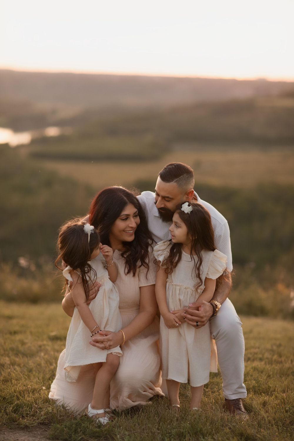 Family of four posing outdoors at golden hour, parents and two young girls in matching cream dresses on a hillside.