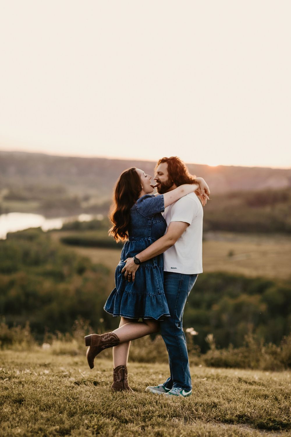Couple embracing at golden hour on a grassy hilltop, woman in denim dress lifted by bearded man with scenic valley view.
