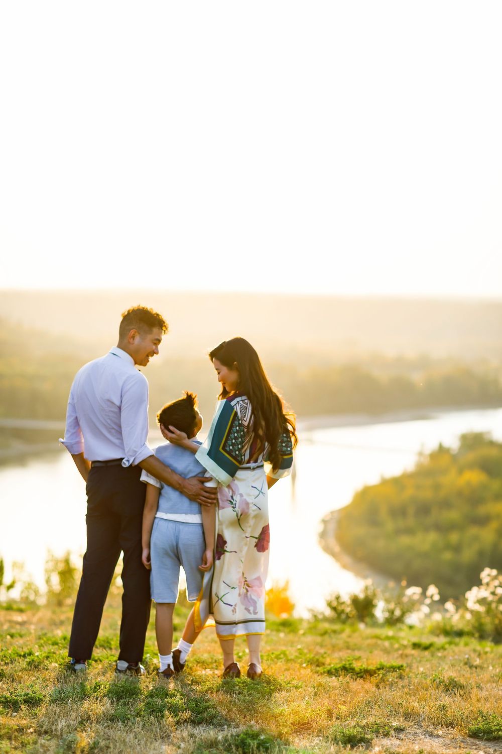 Family of three standing on a hilltop at golden sunset with a scenic river valley in the background.