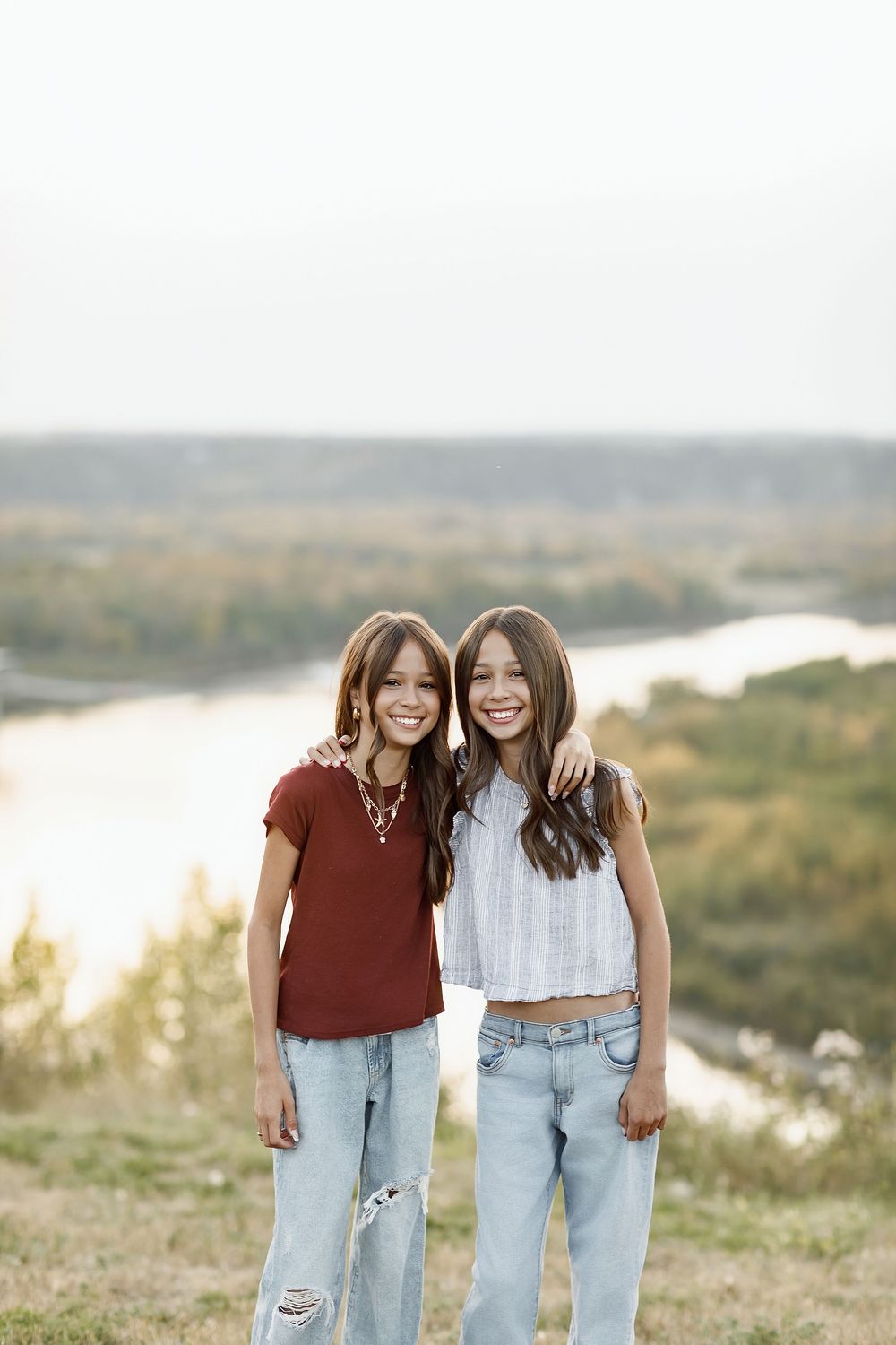 Two smiling teenage girls with brown hair hugging outdoors on a hilltop with a river valley in the background.
